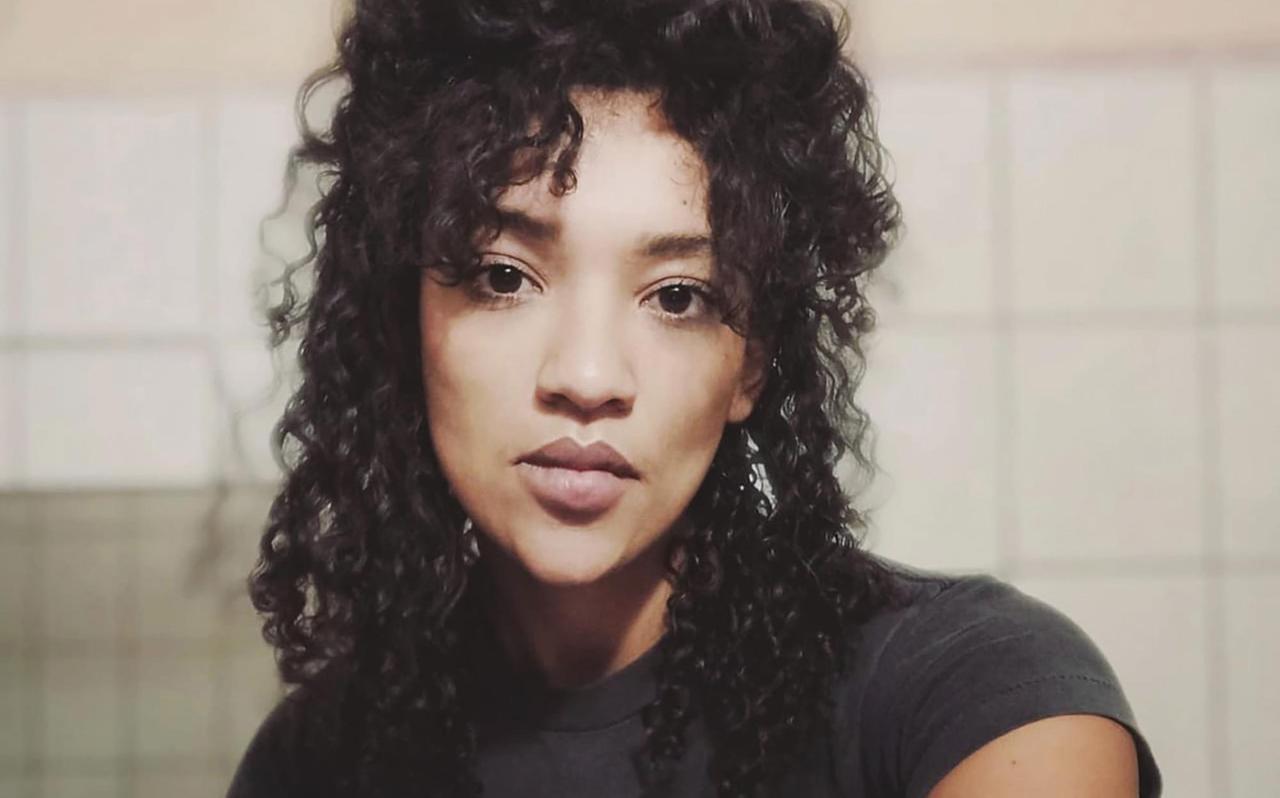 Portrait einer Frau mittleren Alters mit langen, schwarzen Locken. Sie sitzt in einem schwarzen T-Shirt vor einer gekachelten Wand.