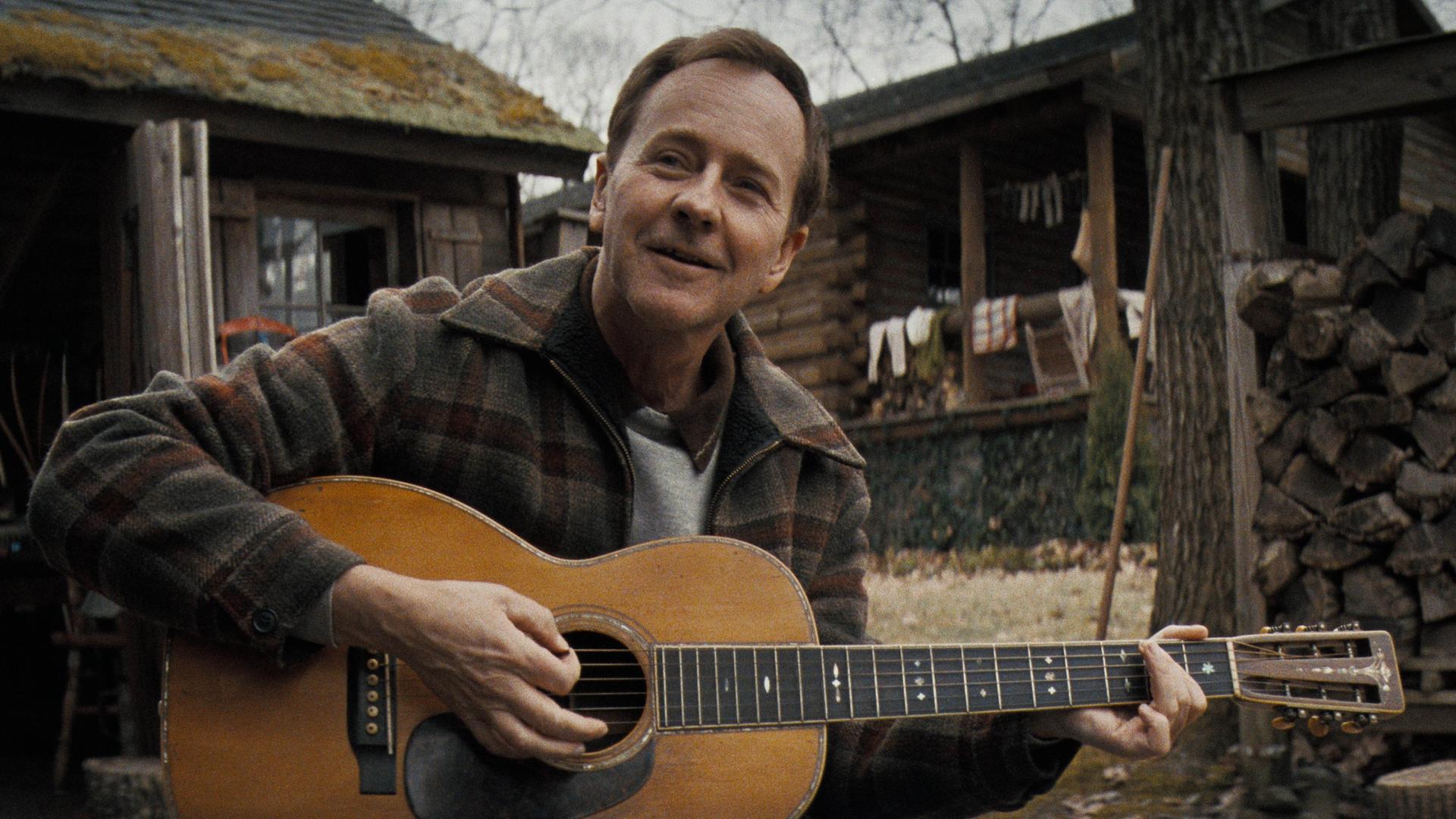 Edward Norton als Folkmusiker Pete Seeger, der vor einer Waldhütte auf einer Gitarre spielt