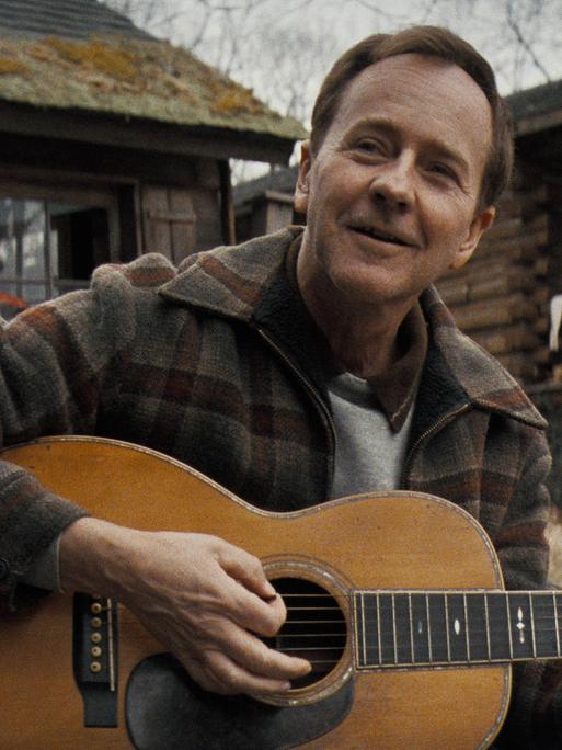 Edward Norton als Folkmusiker Pete Seeger, der vor einer Waldhütte auf einer Gitarre spielt