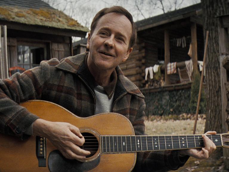 Edward Norton als Folkmusiker Pete Seeger, der vor einer Waldhütte auf einer Gitarre spielt