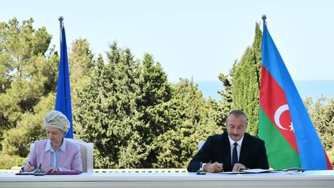 Zwei Personen, eine Frau und ein Mann, sitzen an einem langen weißen Tisch und unterzeichnen Verträge. Im Hintergrund sieht man Bäume, die Flagge der EU und von Aserbaidschan.