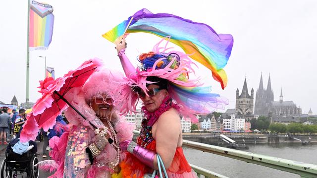 In der Stadt Köln findet eine große CSD-Parade statt. Auf dem Foto tragen zwei Männer bunter Kleider. Einer der Männer hält eine Regenbogen-Flagge hoch. Im Hinter-Grund ist der Kölner Dom zu sehen.