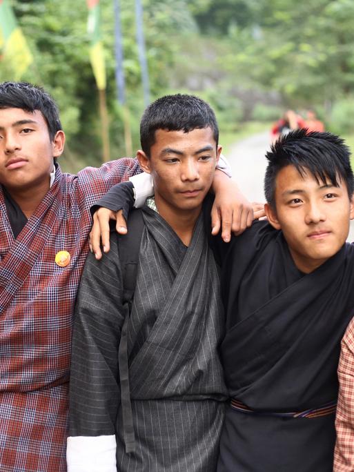Fünf Jugendliche in traditioneller bhutanesischer Kleidung - eine Art karierte Bademäntel - stehen nebeneiner, die Arme umeinander gelegt.