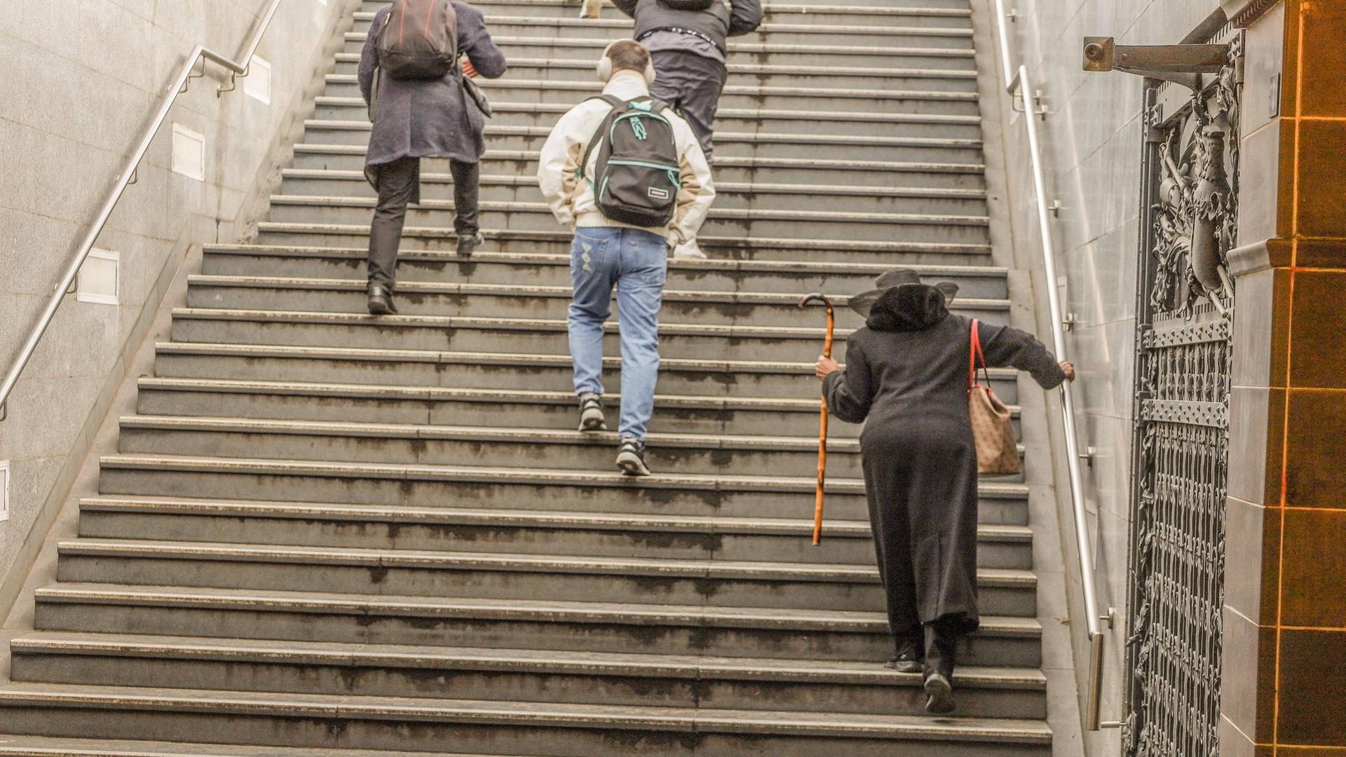 Eine ältere Dame mit Stock und ein junger Mann mit Kopfhörer gehen die Treppe einer U-Bahn herauf. Man sieht sie von hinten.