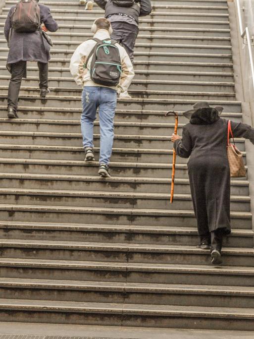 Eine ältere Dame mit Stock und ein junger Mann mit Kopfhörer gehen die Treppe einer U-Bahn herauf. Man sieht sie von hinten.