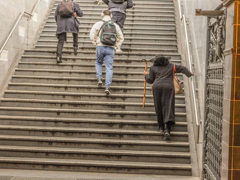 Eine ältere Dame mit Stock und ein junger Mann mit Kopfhörer gehen die Treppe einer U-Bahn herauf. Man sieht sie von hinten.