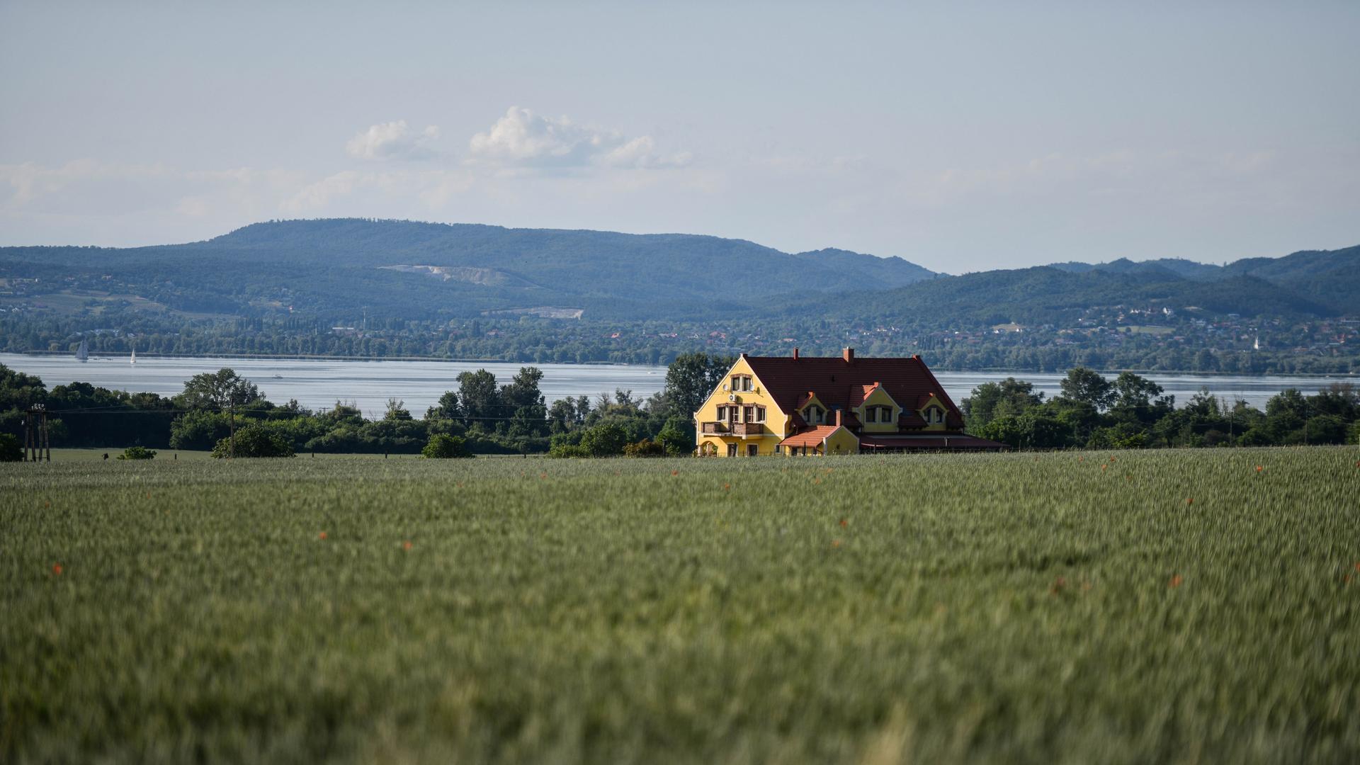Blick auf ein großes Haus in einem Feld, im Hintergrund ist der Plattensee (Balaton) zu sehen. 