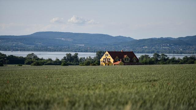 Blick auf ein großes Haus in einem Feld, im Hintergrund ist der Plattensee (Balaton) zu sehen. 