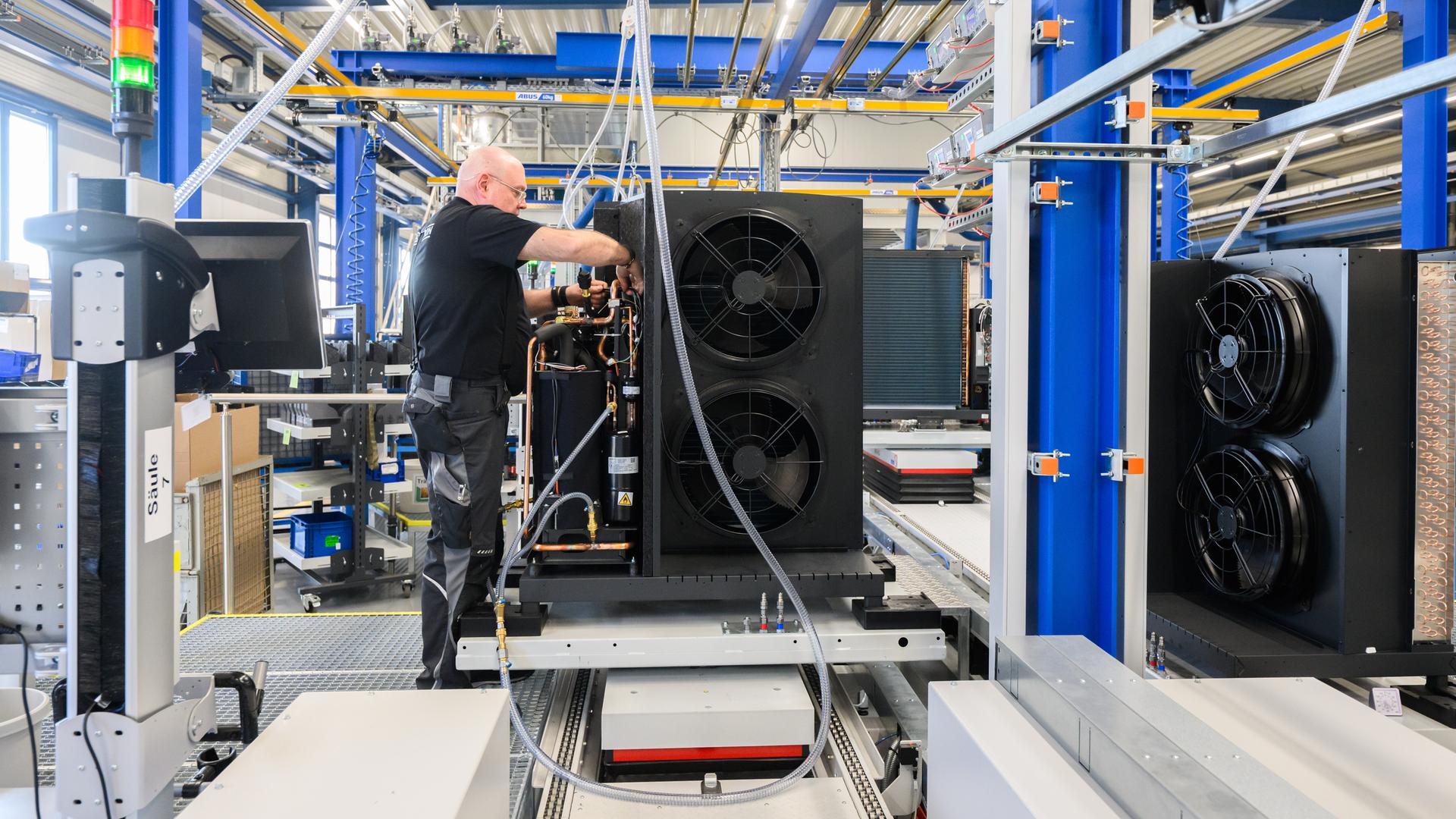 Ein Mitarbeiter arbeitet an einer Wärmepumpe. Zu sehen ist eine Fertigungshalle, blaue Steelen umrahmen das Bild. Ein Mitarbeiter arbeitet an einer Wärmepumpe. Zu sehen ist eine Fertigungshalle, blaue Steelen umrahmen das Bild.