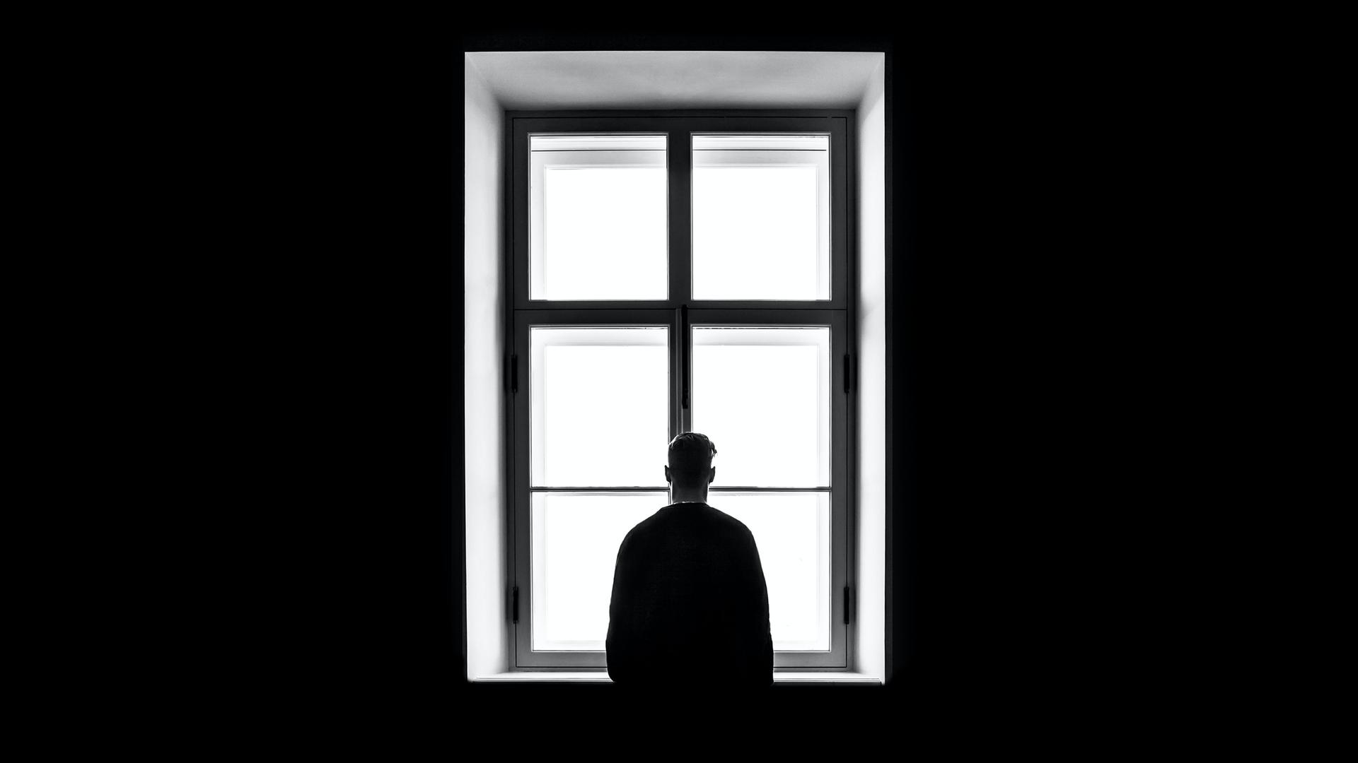 Eine Person steht mit dem Rücken zur Kamera vor einem gleissend hellen Fenster auf diesem schwarz-weiss Bild.