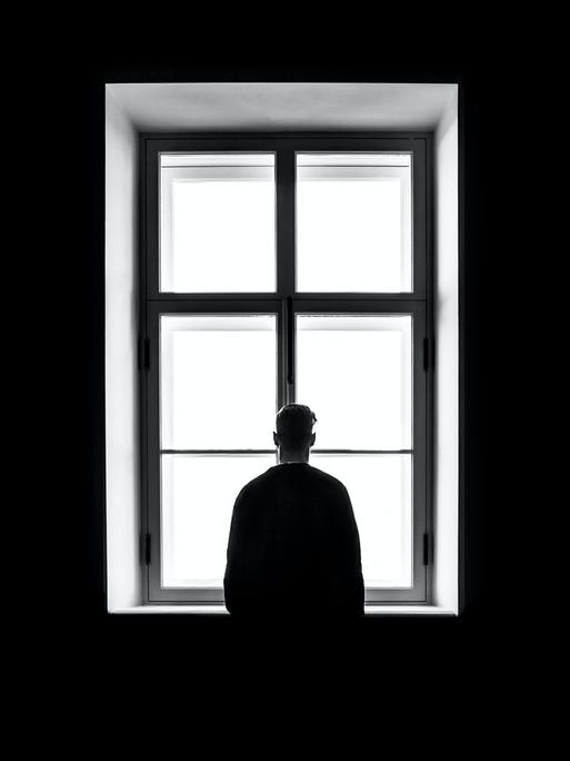 Eine Person steht mit dem Rücken zur Kamera vor einem gleissend hellen Fenster auf diesem schwarz-weiss Bild.