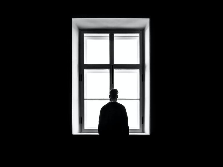 Eine Person steht mit dem Rücken zur Kamera vor einem gleissend hellen Fenster auf diesem schwarz-weiss Bild.