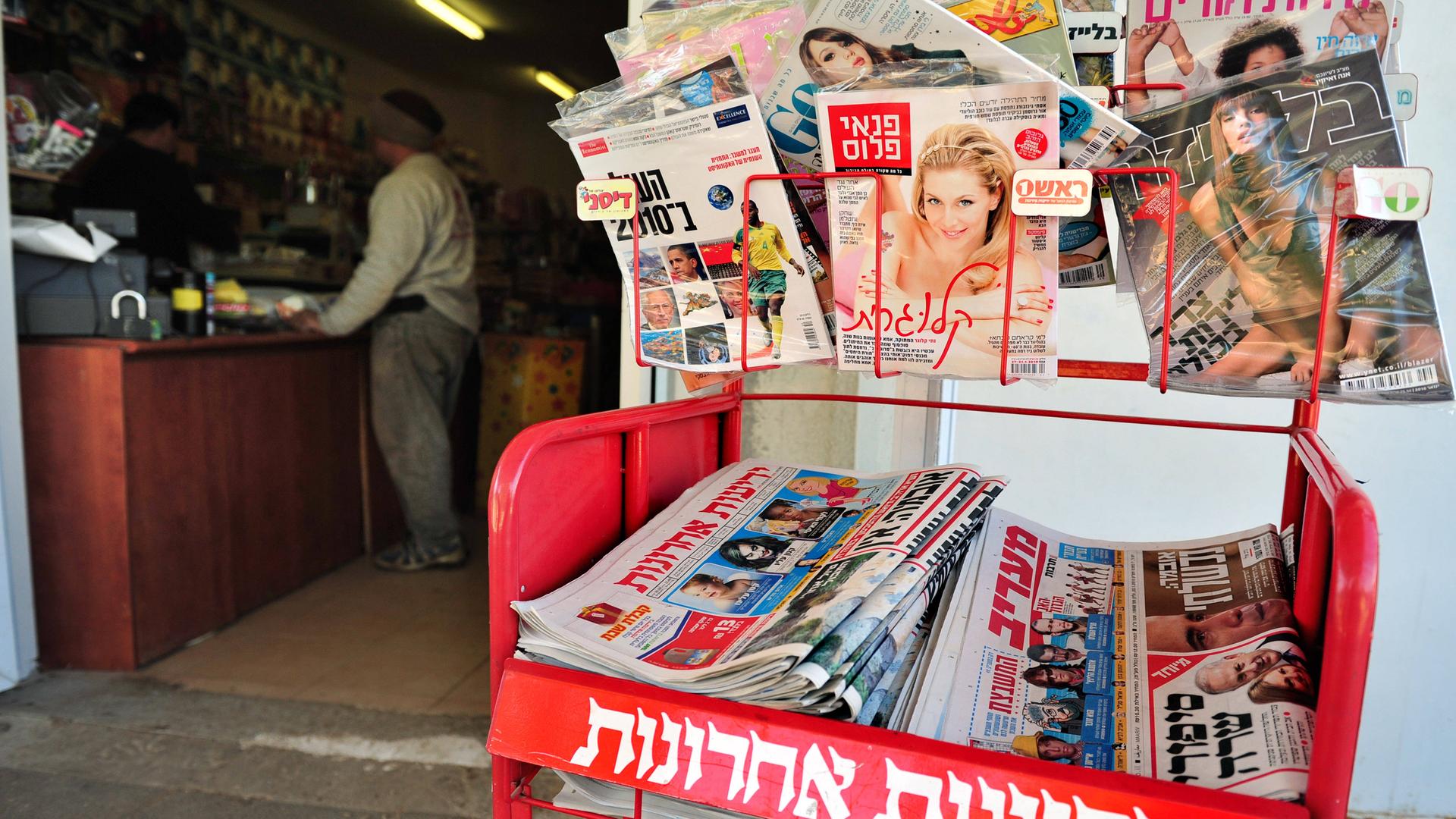 Am Eingang eines Zeitschriftenladens hängen und liegen mehrere Zeitungen. In der linken Bildhälfte sieht man im Innern des Ladens einen Verkäufer und einen Kunden auf beiden Seiten einer Theke. 