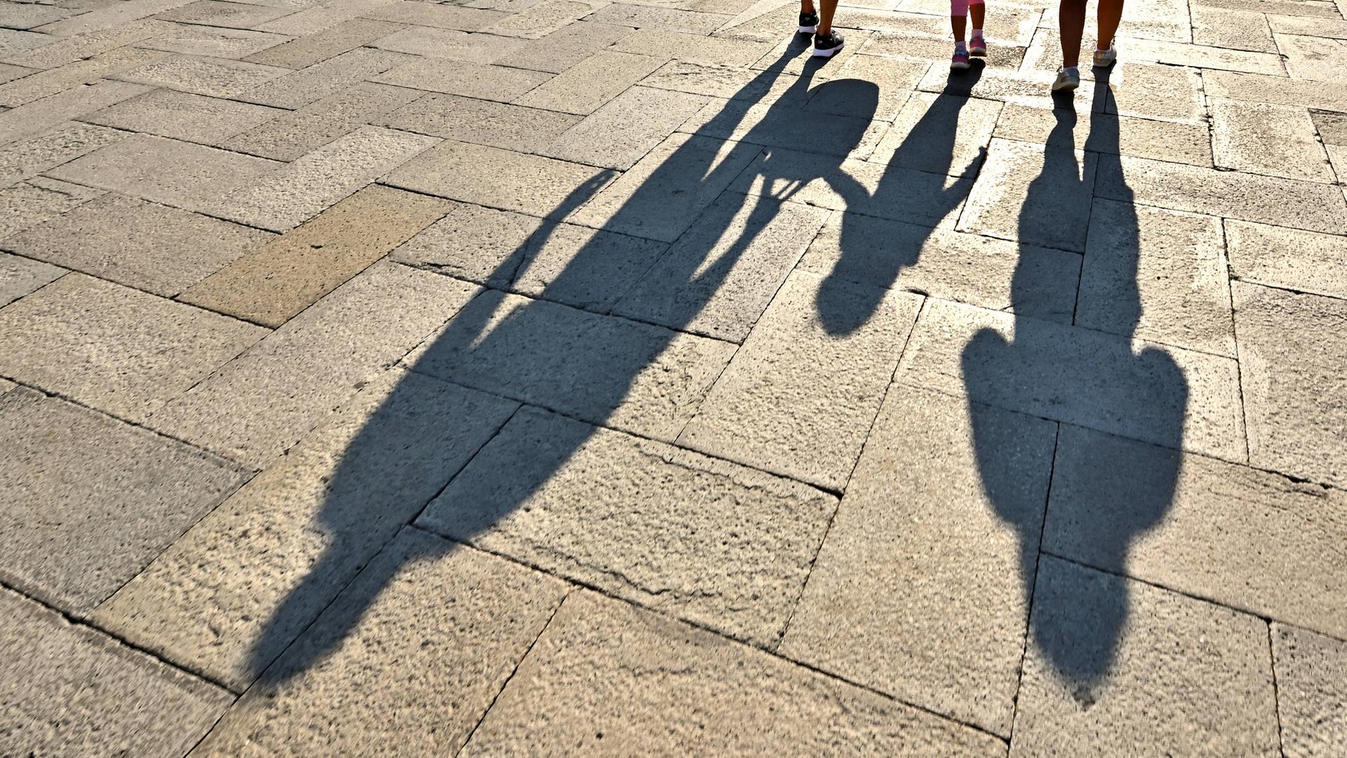 Vater, Mutter und Kind laufen über einen Platz. Ihre Schatten fallen auf den gepflasterten Boden.