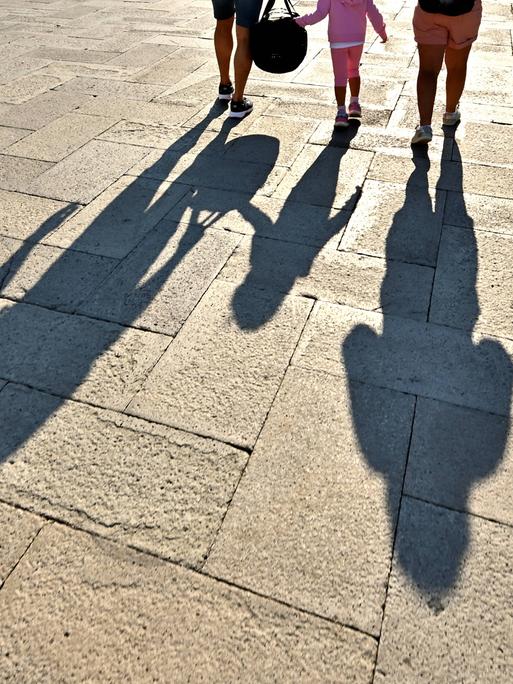 Vater, Mutter und Kind laufen über einen Platz. Ihre Schatten fallen auf den gepflasterten Boden.