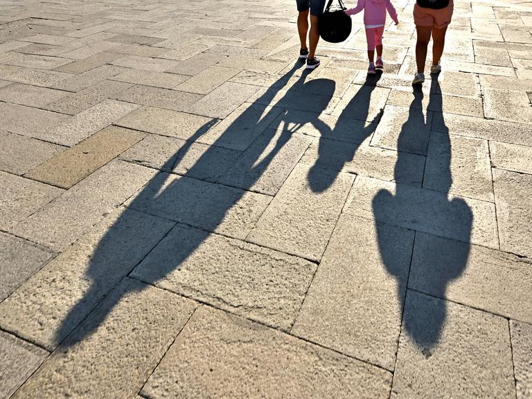 Vater, Mutter und Kind laufen über einen Platz. Ihre Schatten fallen auf den gepflasterten Boden.