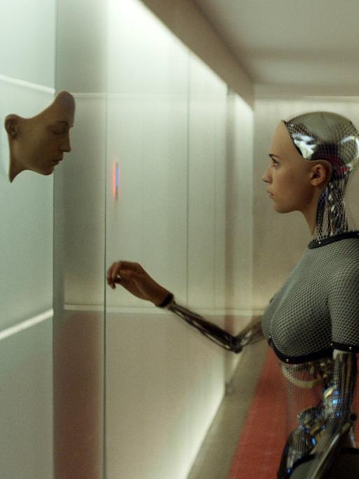 In einem langen Gang mit Betonwänden steht ein weiblich anmutedender Roboter. Die Armgelenke sind als nicht-menschliche Glieder erkennbar. An der linken Wand tausch drei Männer-Gesichter aus dem Beton heraus.