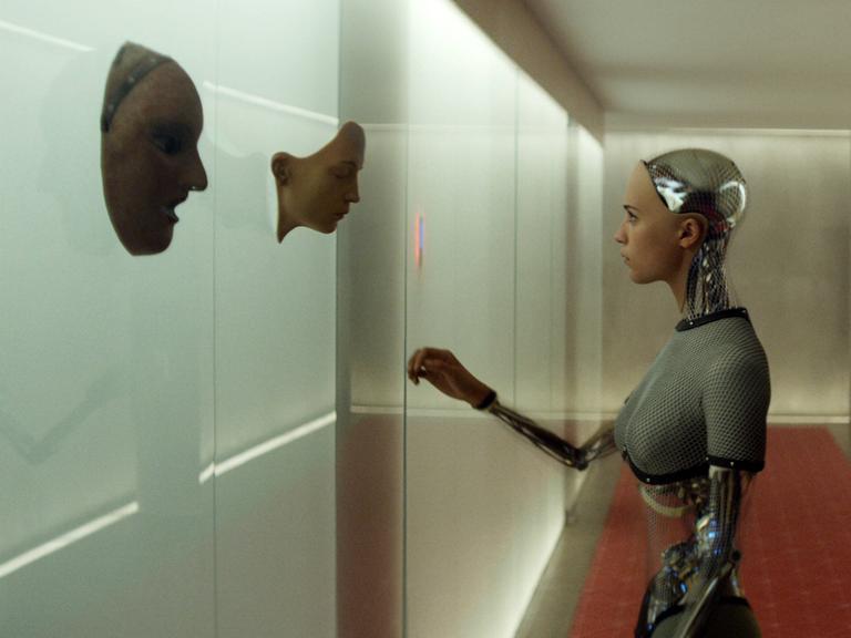 In einem langen Gang mit Betonwänden steht ein weiblich anmutedender Roboter. Die Armgelenke sind als nicht-menschliche Glieder erkennbar. An der linken Wand tausch drei Männer-Gesichter aus dem Beton heraus.