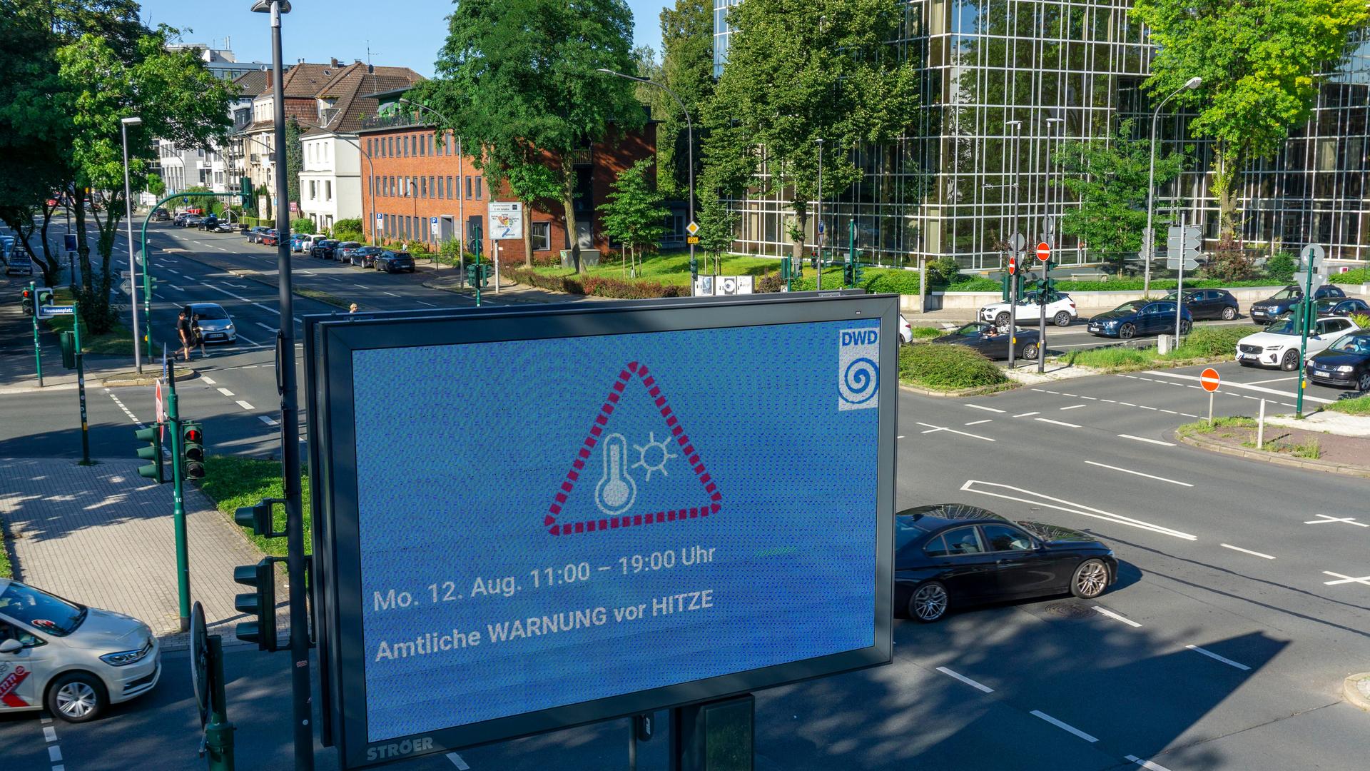 Eine amtliche Warnung vor Hitze steht auf einer digitalen Werbetafel an einer Straßenkreuzung in einer Innenstadt. 