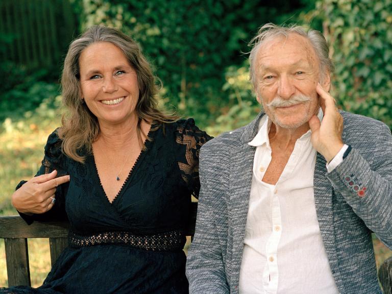 Marie Theres Relin und Franz Xaver Kroetz sitzen lächelnd auf einer Bank im Freien.