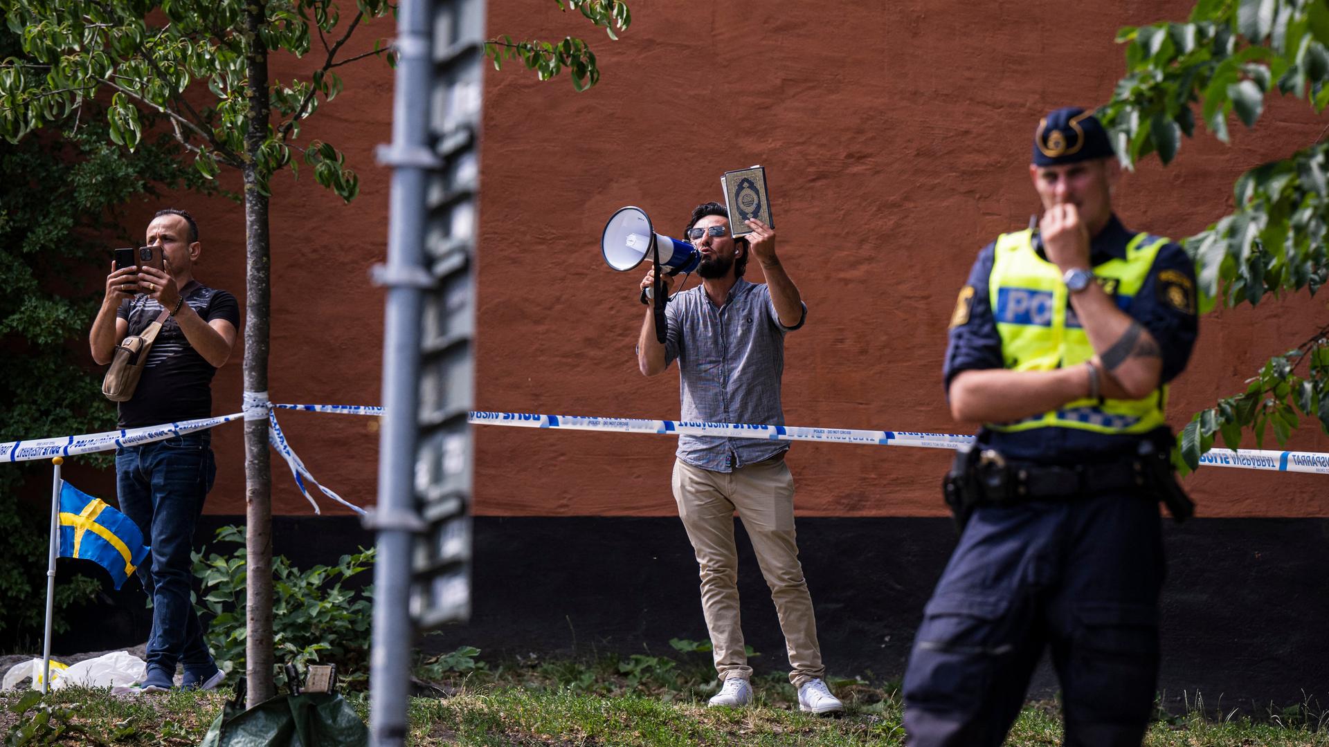 Salwan Momika protestiert außerhalb einer Moschee in Stockholm. Er hält ein Megafon und einen Koran in der Hand.