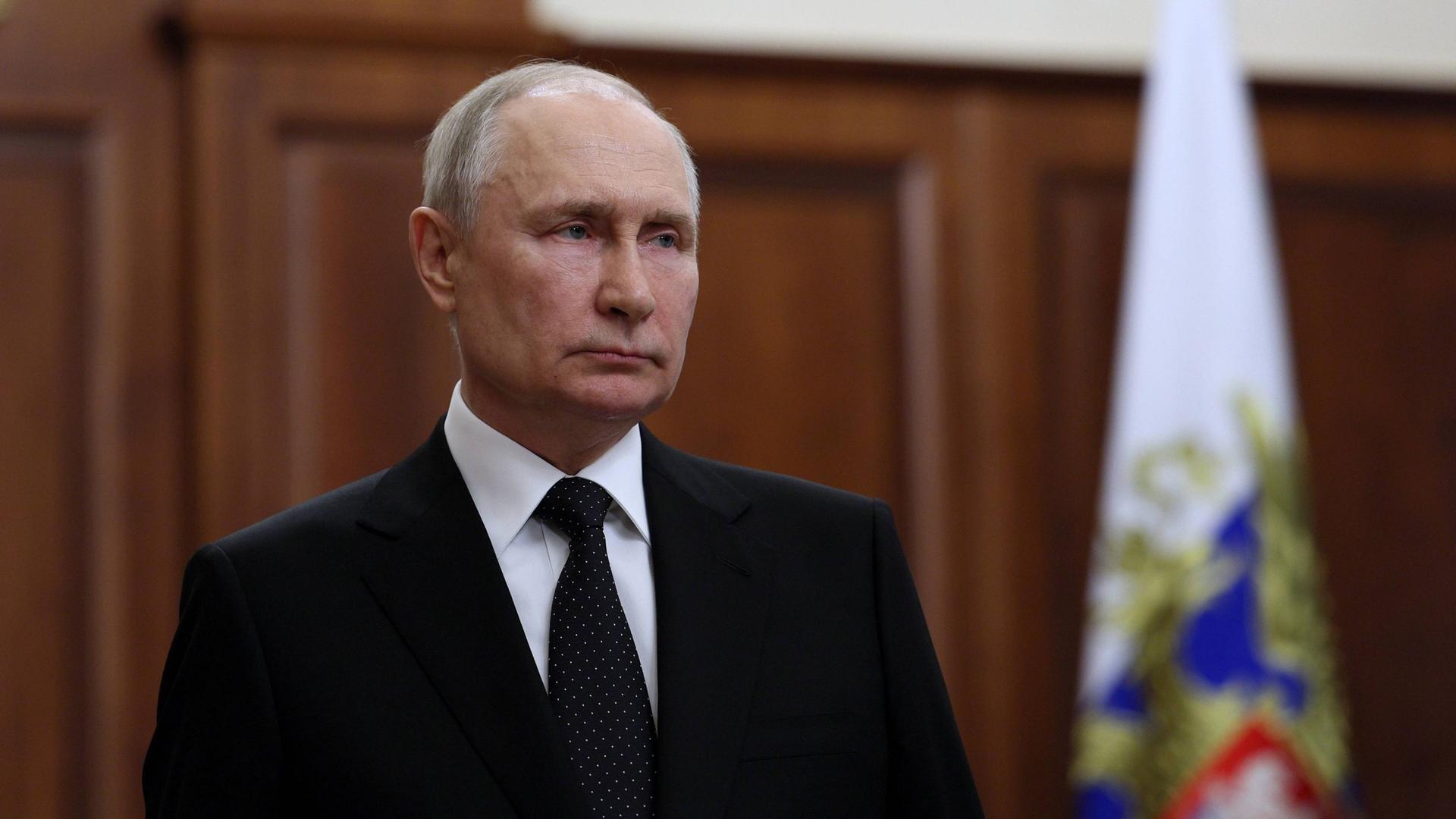 Wladimir Putin steht mit ernstem Blick in einem politisch repräsentativen Raum.
