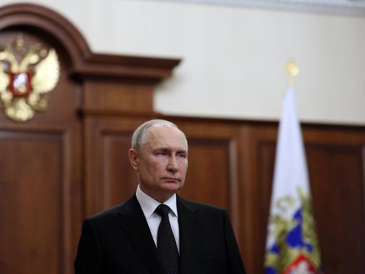 Wladimir Putin steht mit ernstem Blick in einem politisch repräsentativen Raum.