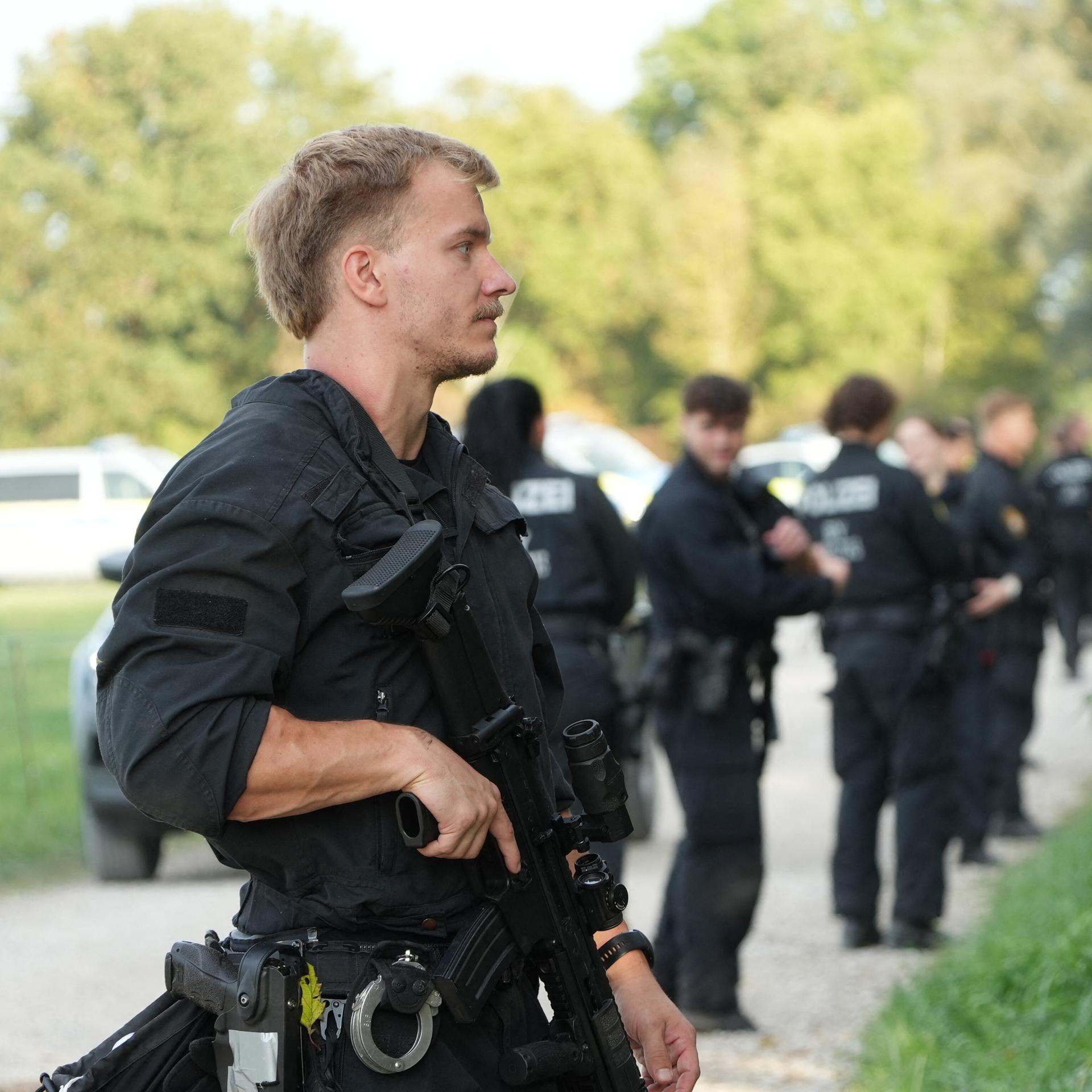 Mehrere Polizisten stehen am Rand eines Waldgebietes. Einer der Polizisten hat eine Waffe im Anschlag.