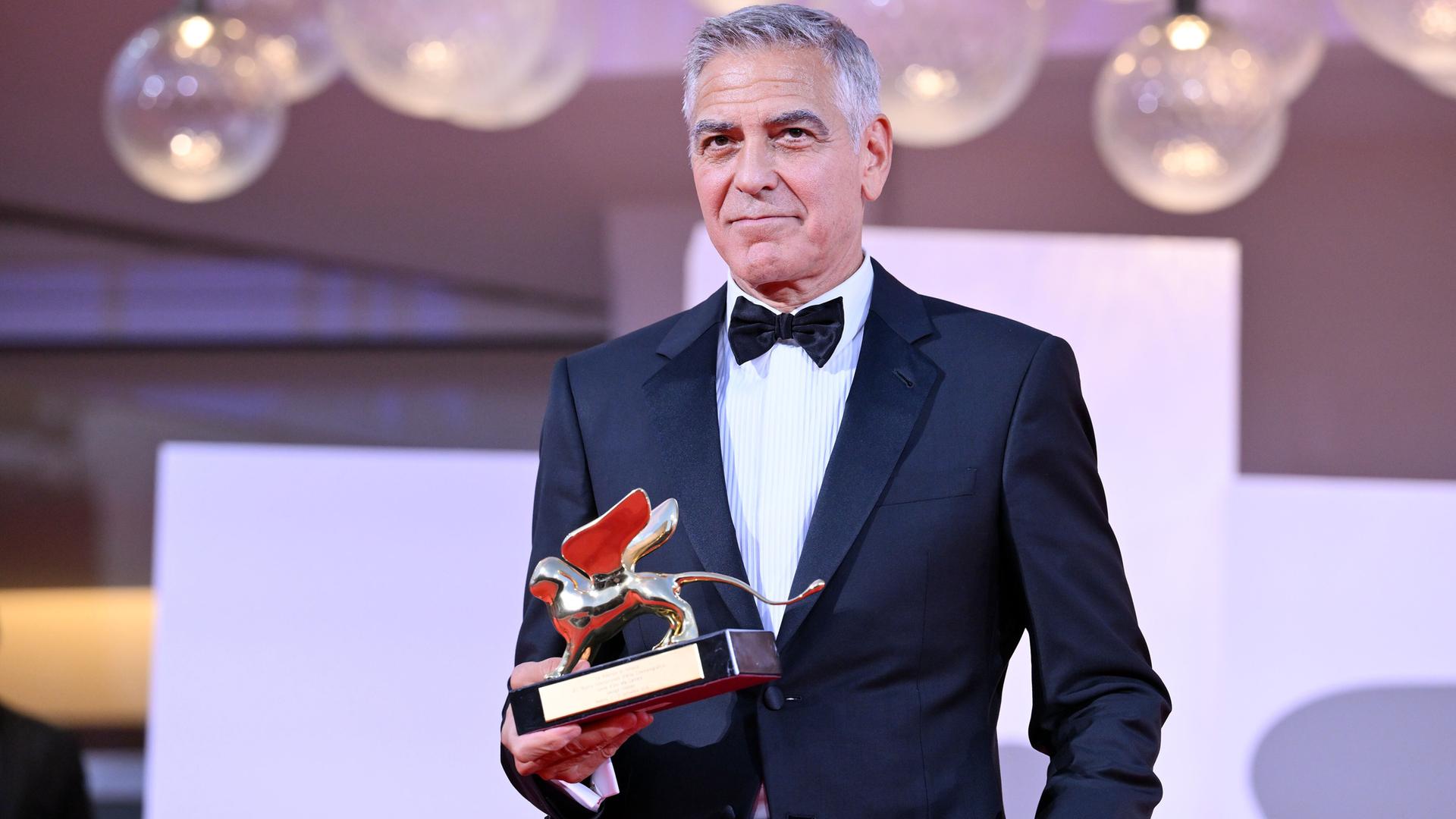 US-Schauspieler George Clooney steht bei den Filmfestspielen in Venedig im eleganten Anzug da. Er hält in einer Hand den Goldenen Löwen, den Ehrenpreis für sein Lebenswerk. 