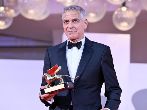 US-Schauspieler George Clooney steht bei den Filmfestspielen in Venedig im eleganten Anzug da. Er hält in einer Hand den Goldenen Löwen, den Ehrenpreis für sein Lebenswerk. US-Schauspieler George Clooney steht bei den Filmfestspielen in Venedig im eleganten Anzug da. Er hält in einer Hand den Goldenen Löwen, den Ehrenpreis für sein Lebenswerk.