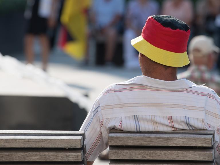 Ein Mann sietzt auf einer Bank und trägt einen Fischerhut in Deutschlandfarben.