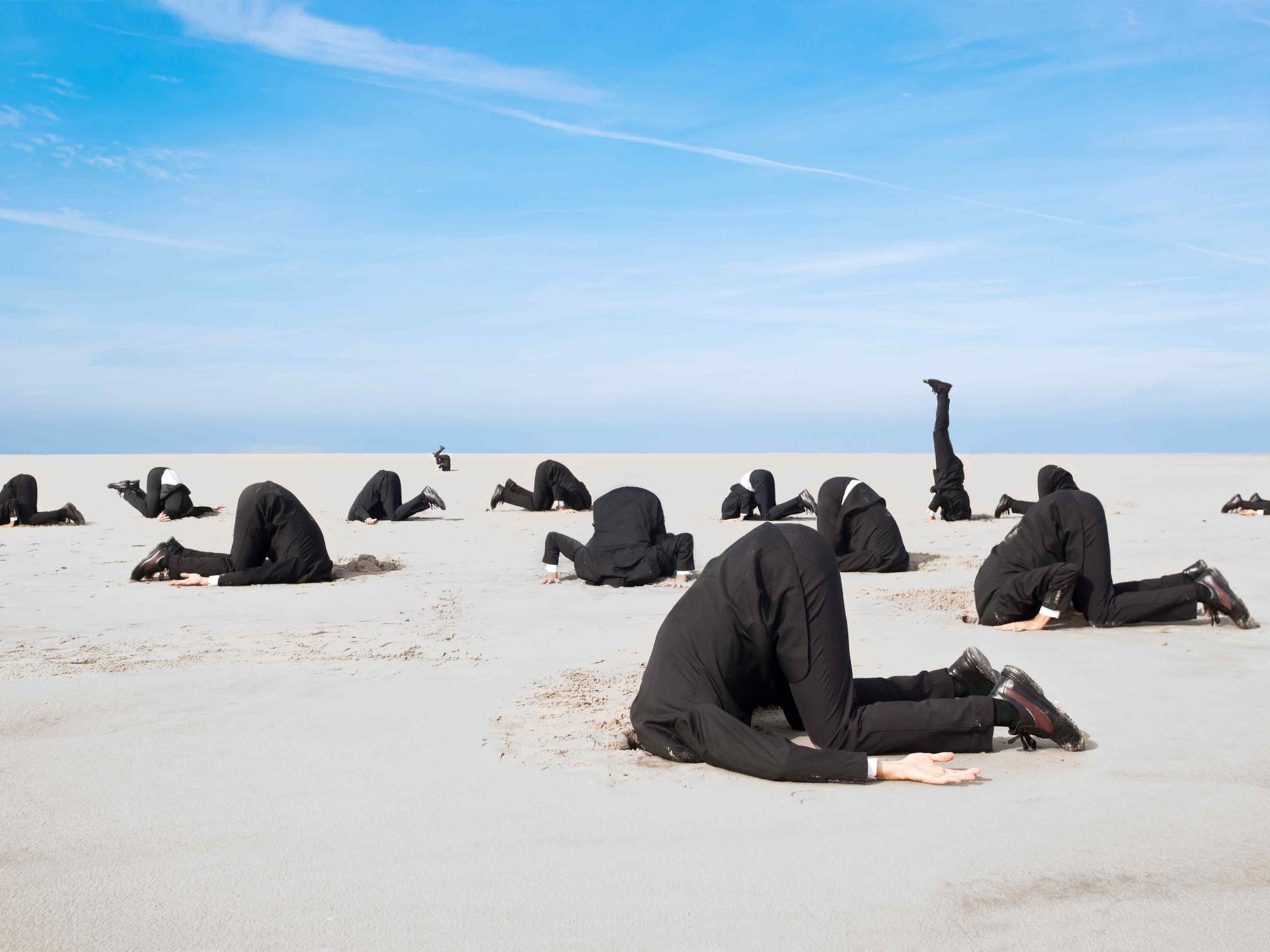 Ein Dutzend Businessmänner in Anzügen stecken vor blauem Himmel die Köpfe in den Sand.