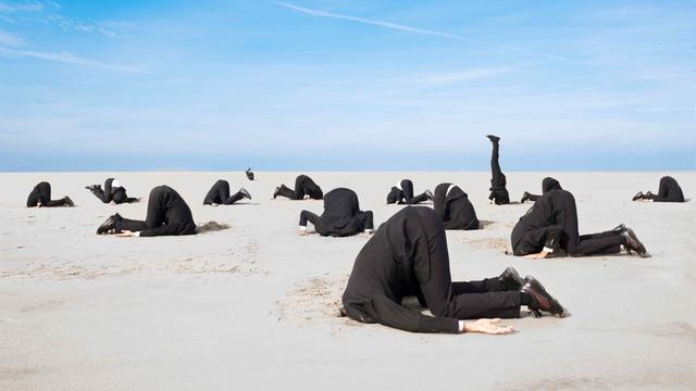 Ein Dutzend Businessmänner in Anzügen stecken vor blauem Himmel die Köpfe in den Sand.