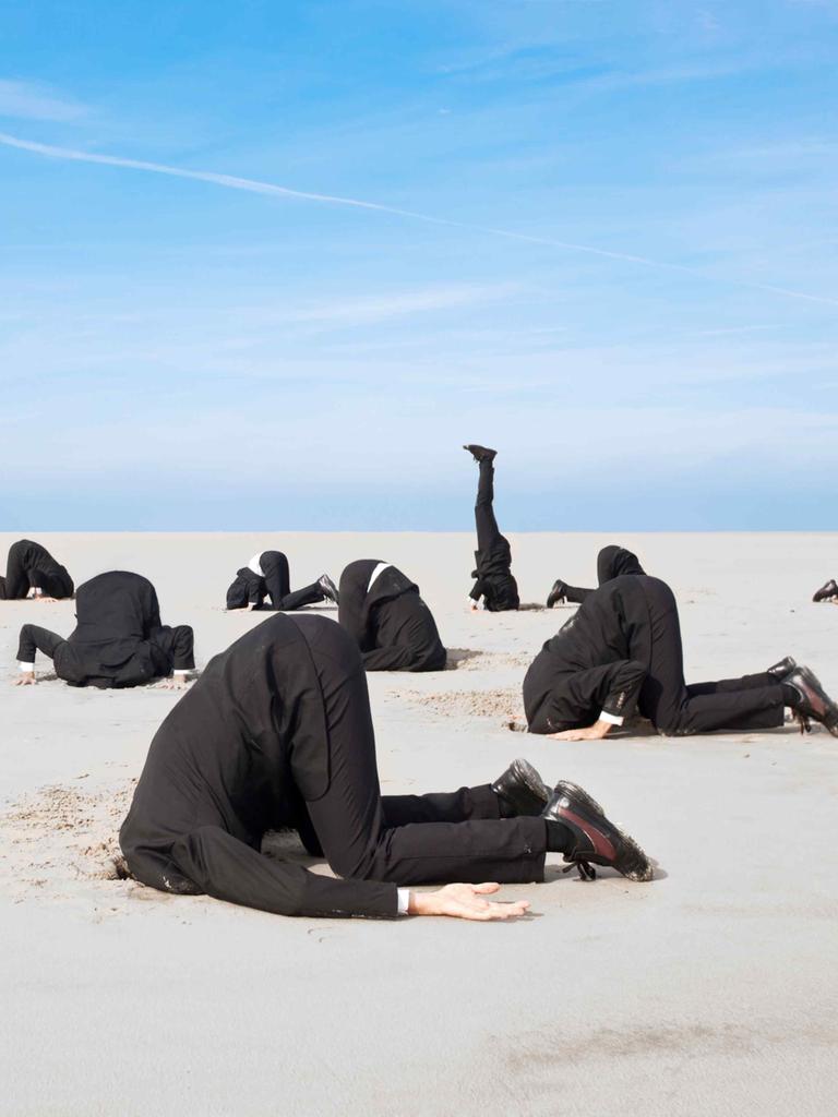 Ein Dutzend Businessmänner in Anzügen stecken vor blauem Himmel die Köpfe in den Sand.