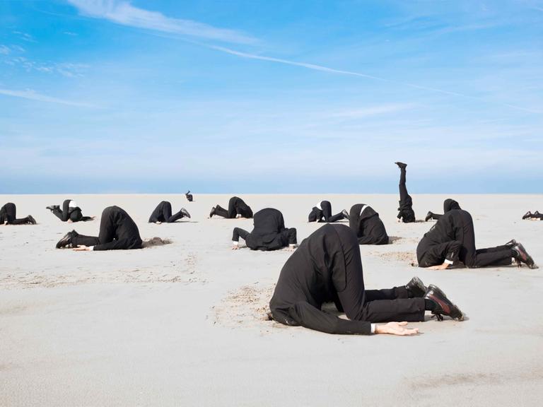 Ein Dutzend Businessmänner in Anzügen stecken vor blauem Himmel die Köpfe in den Sand.