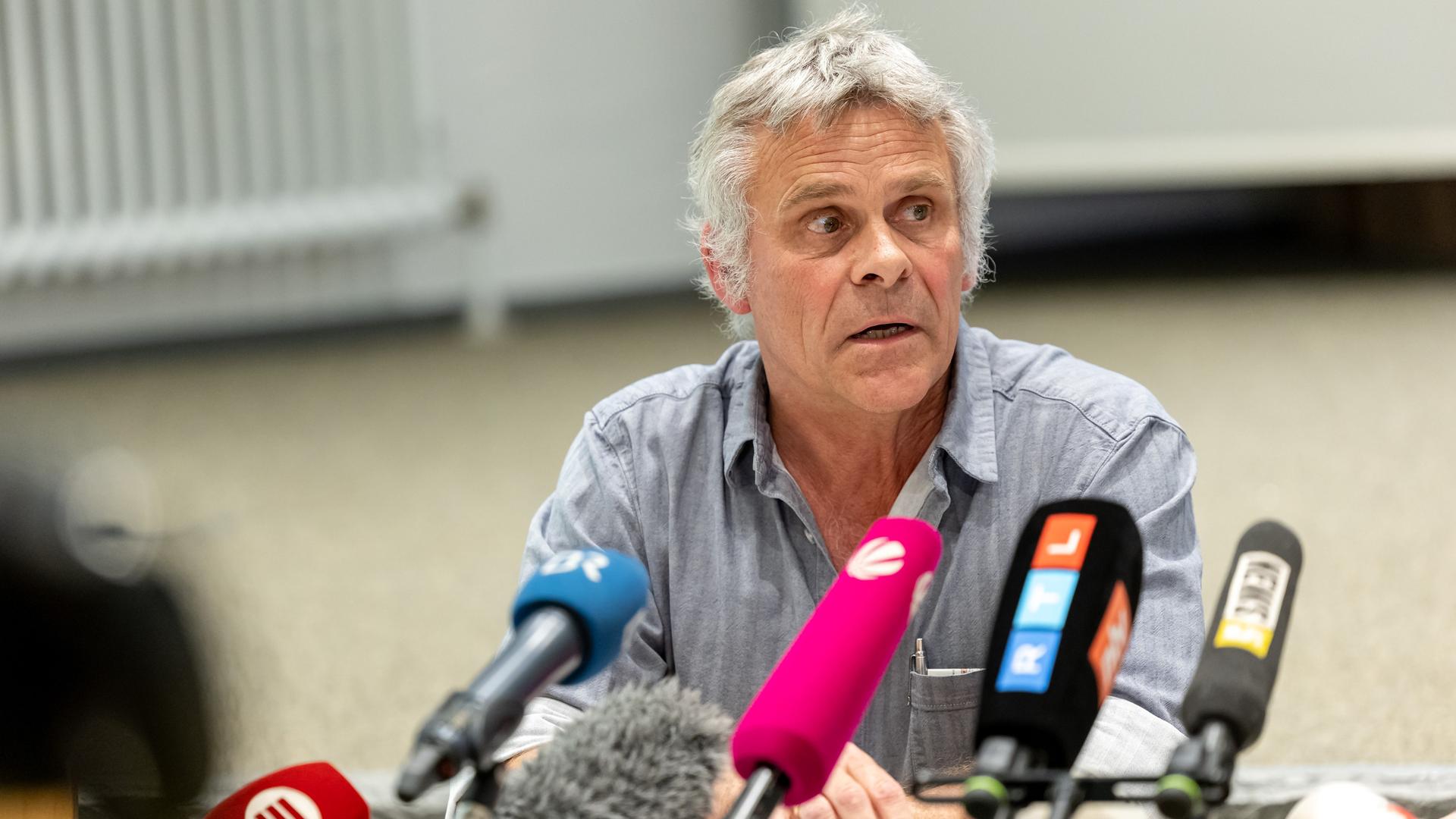 Der grauhaarige Encke sitzt in einem Raum an einem Tisch und spricht in mehrere Mikrofone von Sendern. Im Hintergrund unscharf eine Wand mit Heizung.