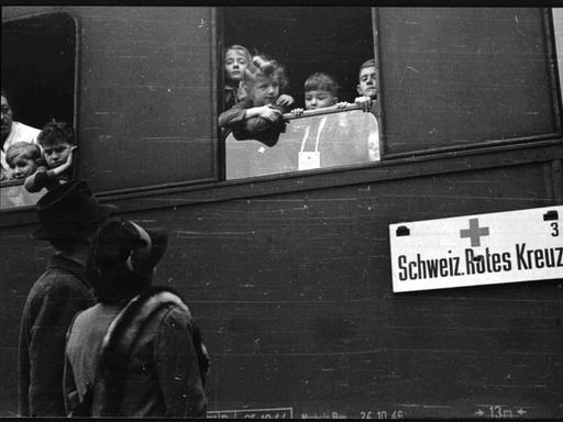 Schwarz-weiß-Foto: Kinder schauen aus einem Zugfenster. Am Zug ist ein Schild befestigt: "Schweizerisches Rotes Kreuz".