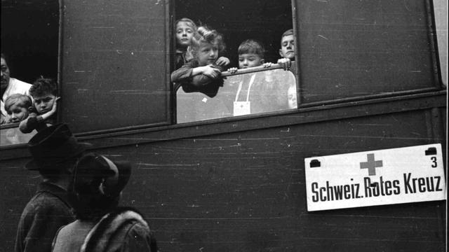 Schwarz-weiß-Foto: Kinder schauen aus einem Zugfenster. Am Zug ist ein Schild befestigt: "Schweizerisches Rotes Kreuz".