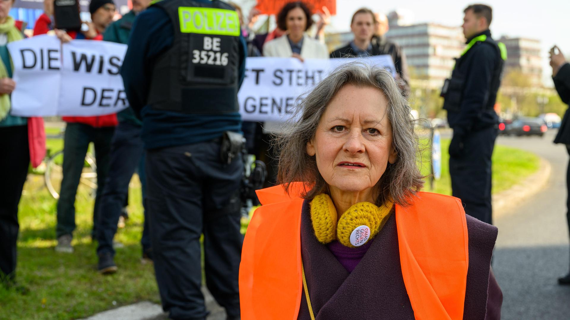 Marion Fabian (72, Künstlerin), im Hintergrund Polizei und sich solidarisierende Wissenschaftler bei der Blockade der Letzten Generation am Ernst-Reuter-Platz, Berlin, 28.04.23