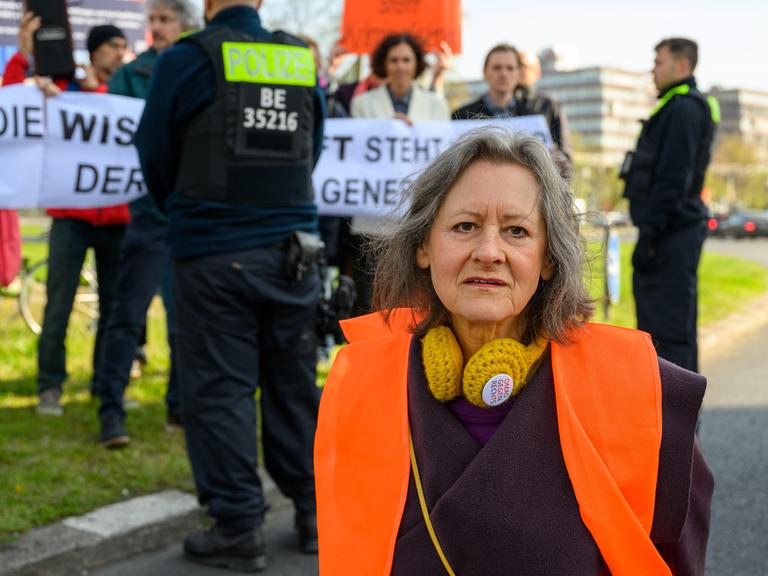 Marion Fabian (72, Künstlerin), im Hintergrund Polizei und sich solidarisierende Wissenschaftler bei der Blockade der Letzten Generation am Ernst-Reuter-Platz, Berlin, 28.04.23