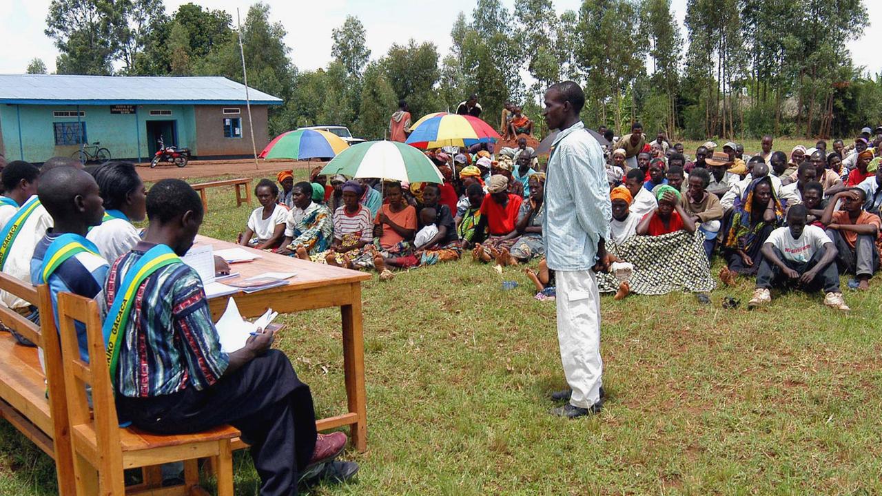 Ein Mann steht auf einer Wiese in Ruanda vor einem traditionellen Dorfgericht. An einem Tisch sitzen die Richter und auf der Wiese Zuschauer. Ein Mann steht auf einer Wiese in Ruanda vor einem traditionellen Dorfgericht. An einem Tisch sitzen die Richter und auf der Wiese Zuschauer.
