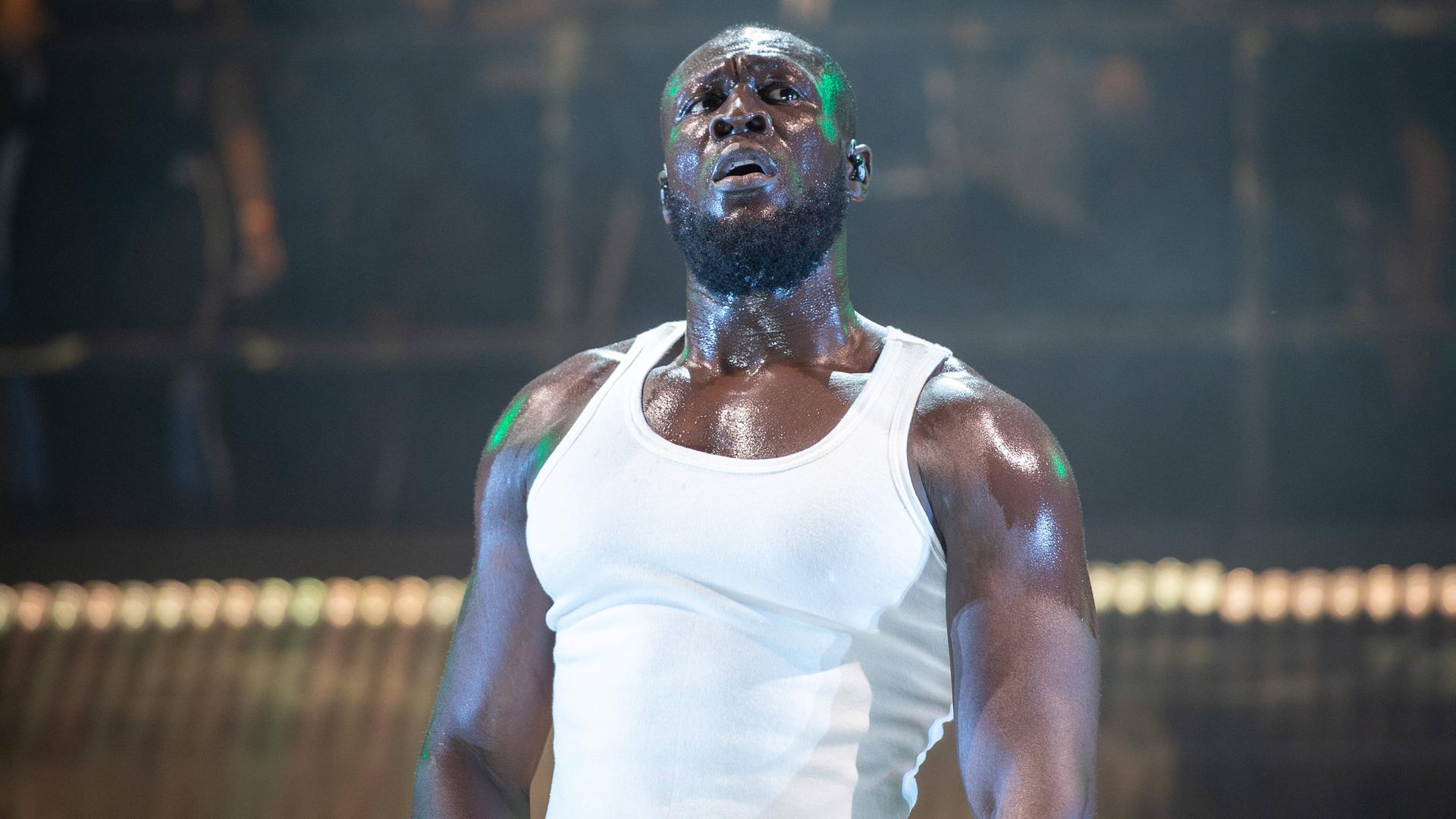 Musiker Stormzy steht im weißen Muscle-Shirt auf der Bühne. Sein Körper glänzt, Schweißperlen sind zu sehen. Musiker Stormzy steht im weißen Muscle-Shirt auf der Bühne. Sein Körper glänzt, Schweißperlen sind zu sehen.