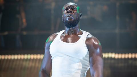 Musiker Stormzy steht im weißen Muscle-Shirt auf der Bühne. Sein Körper glänzt, Schweißperlen sind zu sehen.