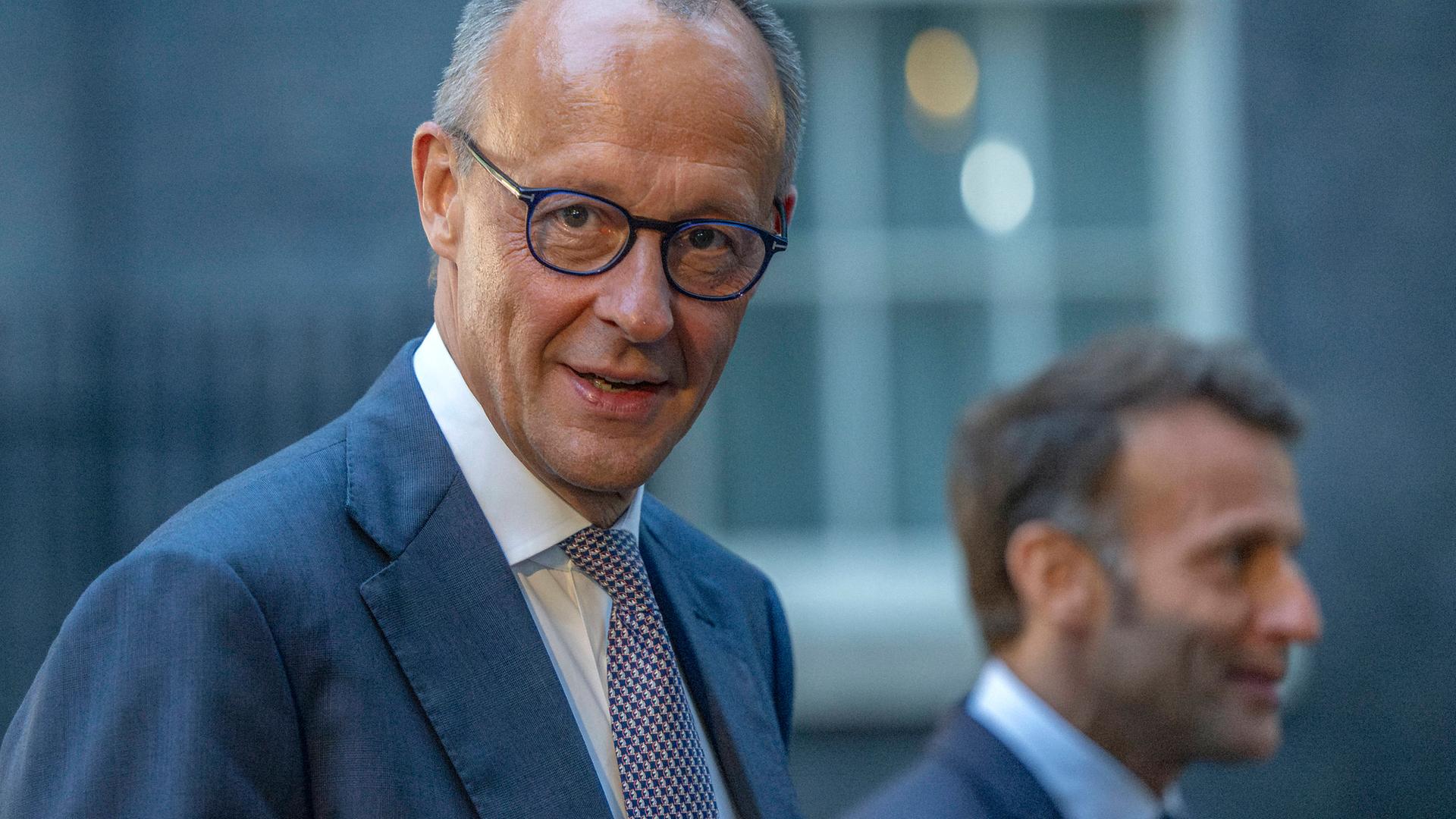 Kanzler Friedrich Merz schaut direkt in die Kamera, unscharf im Hintergrund Frankreichs Präsident Macron beim Ukraine-Gipfel in London am 7. Juni 2026