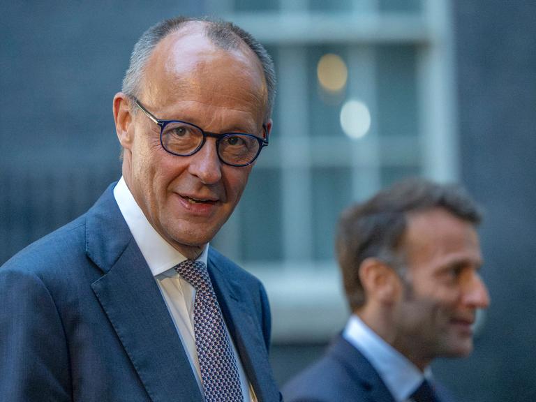 Kanzler Friedrich Merz schaut direkt in die Kamera, unscharf im Hintergrund Frankreichs Präsident Macron beim Ukraine-Gipfel in London am 7. Juni 2026