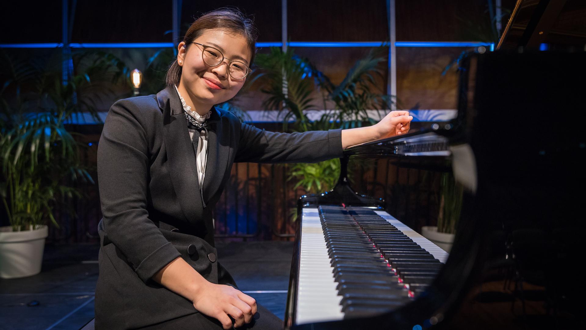 Yumeka Nakagawa sitzt mit ihrer Brille und ihren nach hinten gebundenen Haaren offen lachend am Klavier.