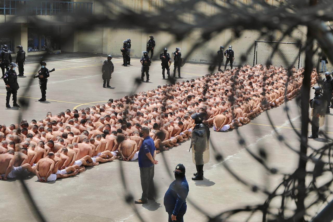 Gefängnis in El Salvador. Menschen sitzen auf dem Boden, Wärter stehen mit Maschinengewehren daneben.