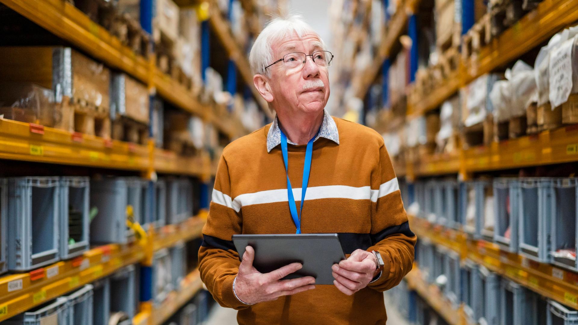 Ein älterer Mann steht mit einem Tablet in der Hand im Gang eines Warenlagers.