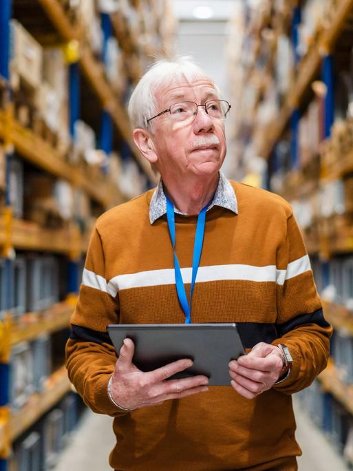 Ein älterer Mann steht mit einem Tablet in der Hand im Gang eines Warenlagers.