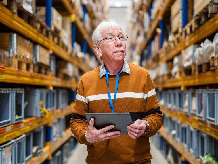 Ein älterer Mann steht mit einem Tablet in der Hand im Gang eines Warenlagers.